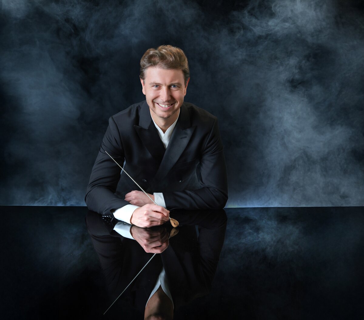 Conductor Jayce Ogren smiling and holding a baton in a studio portrait